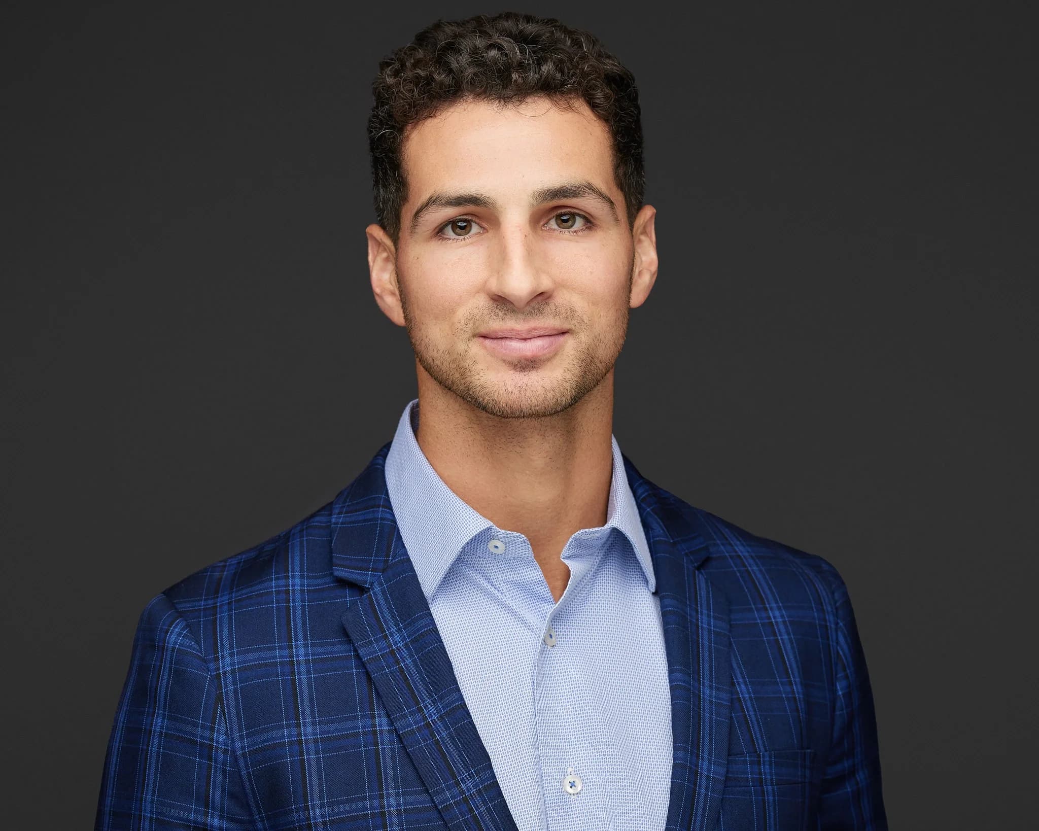 Professional headshot of a man with curly dark hair, wearing a blue plaid blazer over a light blue shirt, against a dark background.