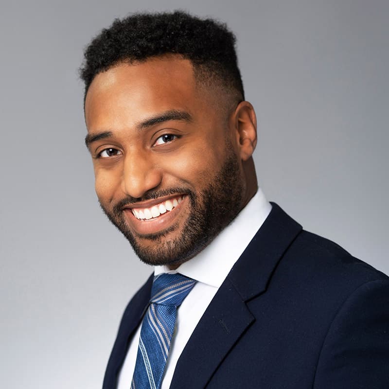 Professional headshot of a smiling man in a navy suit and striped tie against a gray background