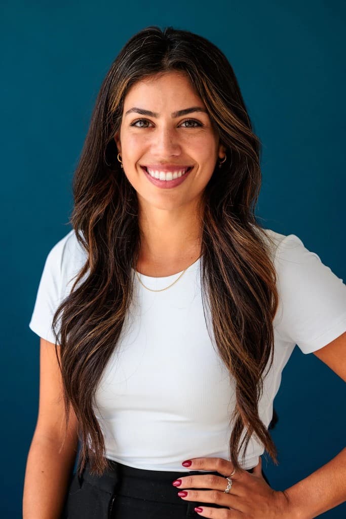 Professional headshot of a smiling woman with long dark hair in a white t-shirt against a teal background