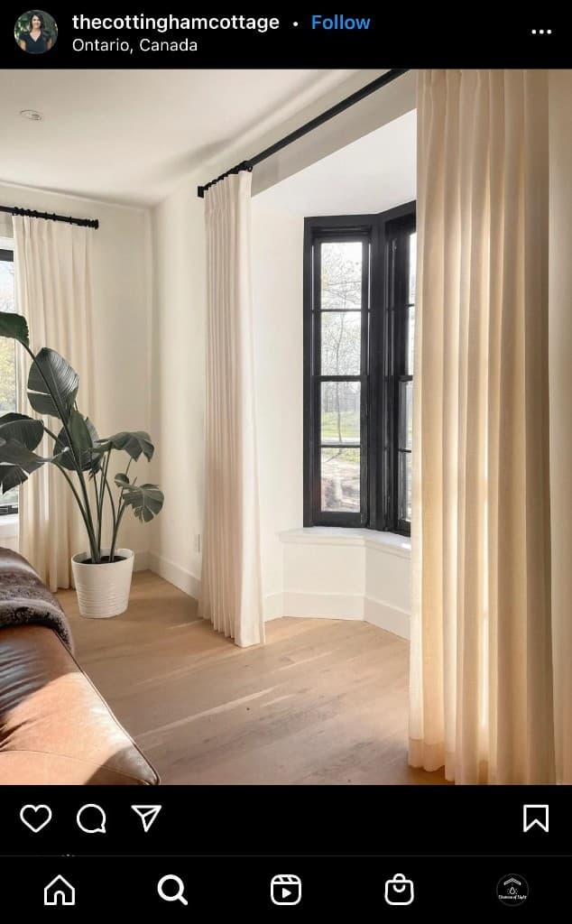 Bright living room with black-framed bay window, cream curtains, and a large potted plant