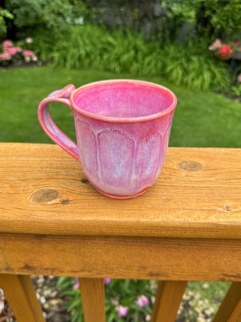 Pink handmade ceramic mug with carved petal details and curved handle on a wooden deck rail